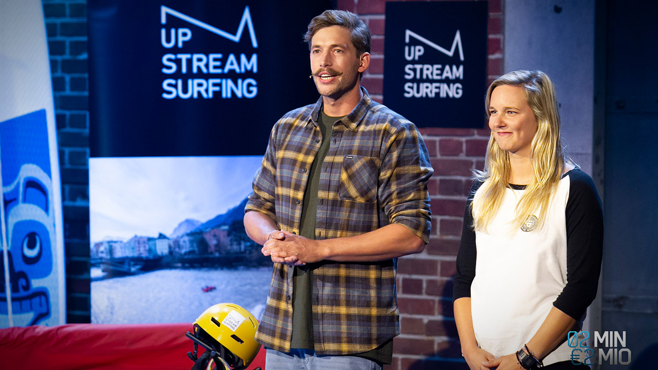 Michael und Livia halten den Pitch. Im Hintergrund sieht man das Logo von UP STREAM SURFING, ein stylisierten Berg