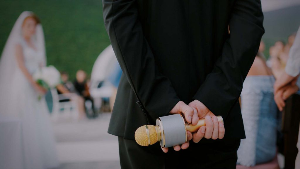 Trauzeuge mit Mikrofon in den Händen bei Outdoor-Hochzeit – bereit für humorvolle Hochzeitsrede vor Braut und Gästen im Hintergrund