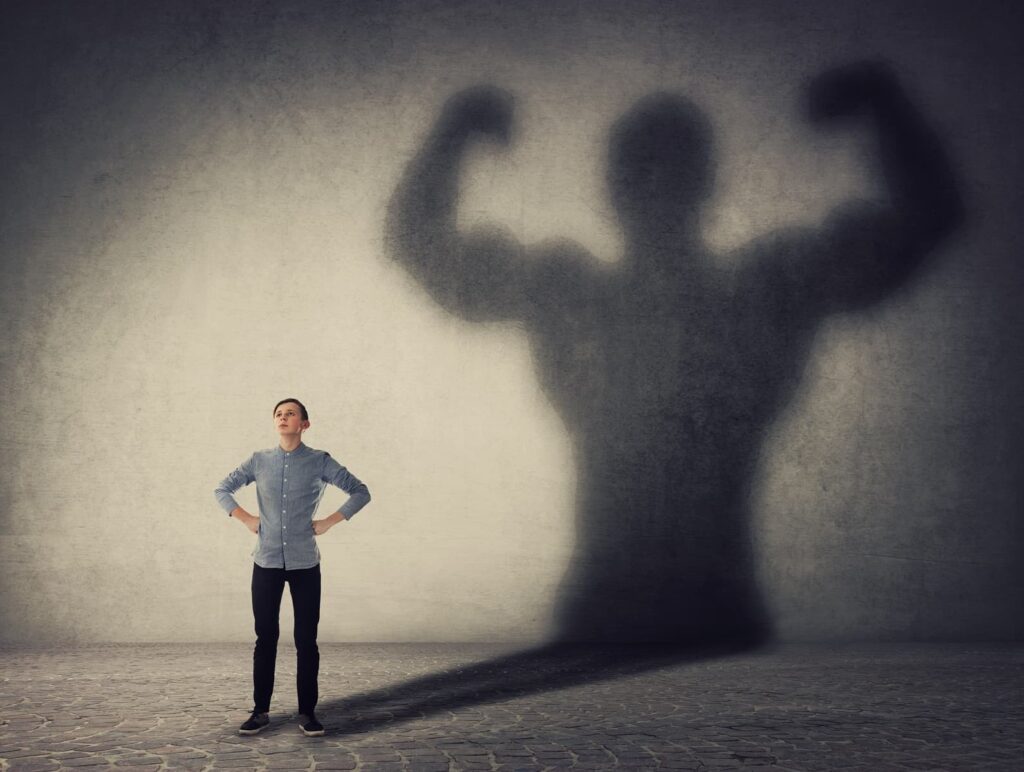 Junger Mann mit selbstbewusster Haltung, dessen Schatten an der Wand einen starken, muskulösen Körper zeigt – Symbolbild für die positiven Effekte von Power Posing