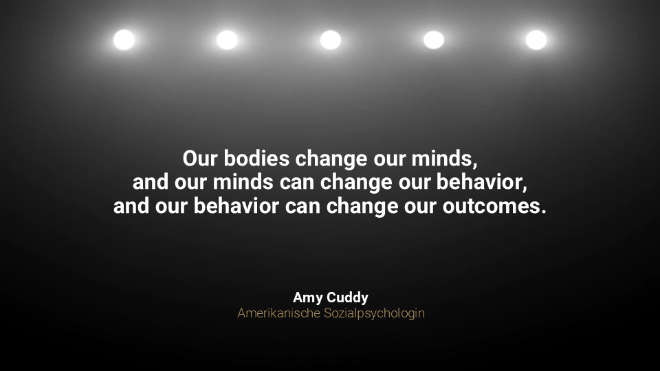 Zitat von Amy Cuddy auf dunklem Bühnenhintergrund mit Scheinwerfern: „Our bodies change our minds, and our minds can change our behavior, and our behavior can change our outcomes.“ – Aussage zur Verbindung von Körperhaltung, Denken und Wirkung