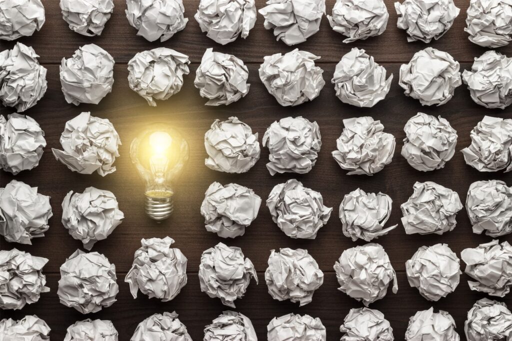 Symbolbild für den kreativen Story-Finder-Prozess: Eine leuchtende Glühbirne zwischen vielen zerknüllten Papierkugeln steht für die eine zündende Story-Idee inmitten unzähliger verworfener Gedanken.